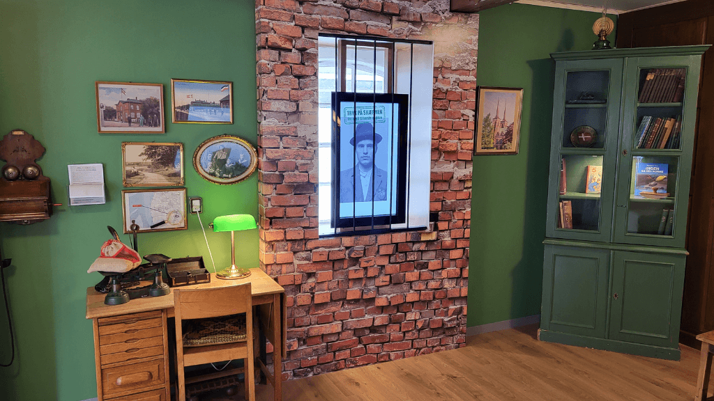 Office with green walls, a brick wall in the middle and a desk with a wooden chair, and a green display cabinet